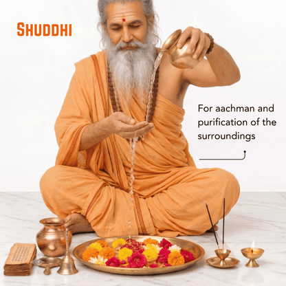 A man dressed as a Shuddhi sadhu sits cross-legged on a marble floor, pouring sacred water from a brass pot into a cup. He wears an orange robe and has a grey beard.
