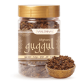 A jar of Vasudham Afghan guggul herbal powder with a gold lid and brown label, shown alongside a small wooden bowl filled with the dark brown powder on a white background.