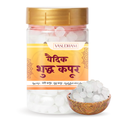 A clear plastic jar of Vasudham pure Kapur tablets with a gold lid and orange label, shown alongside a small wooden bowl filled with the white, translucent crystals.