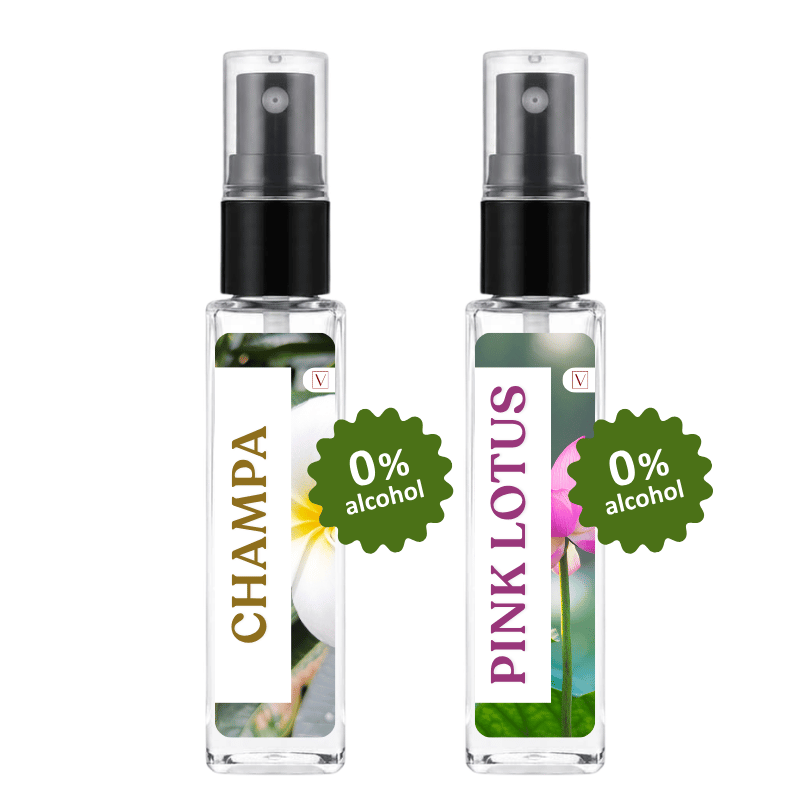 Two clear glass perfume spray bottles with black atomizers on a white background. The left bottle has a white label with gold 'CHAMPA' text and a white flower photo, with a green '0% alcohol' badge.
