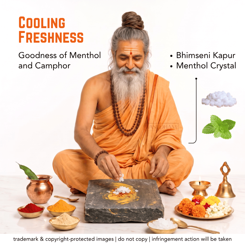 A man with a long gray beard and hair in a bun, dressed in orange robes, sits cross-legged on a white background while preparing a paste on a dark stone slab. Surrounding him are small bowls of colorful powders, a copper pot with a leaf, a brass lamp, and a plate of marigolds, flowers, and crystals.