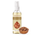 A 100 ml bottle of Vasudham Chandan Jal Premium Sandal Water, featuring a white spray cap and a beige label with a brown wood-grain design and a teardrop-shaped image of sandal wood chips. A small wooden bowl filled with sandal wood chips sits next to the bottle on a clean white background.