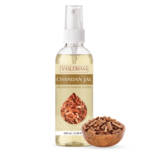 A 100 ml bottle of Vasudham Chandan Jal Premium Sandal Water, featuring a white spray cap and a beige label with a brown wood-grain design and a teardrop-shaped image of sandal wood chips. A small wooden bowl filled with sandal wood chips sits next to the bottle on a clean white background.