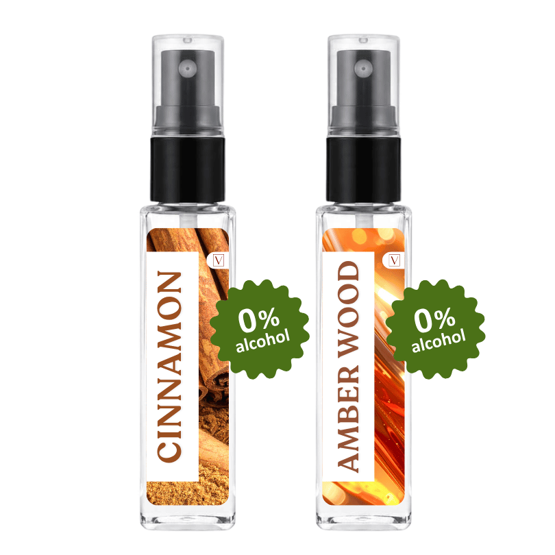Two clear glass perfume spray bottles on a white background, labeled Cinnamon and Amber Wood, each with a black spray top and a green '0% alcohol' badge.