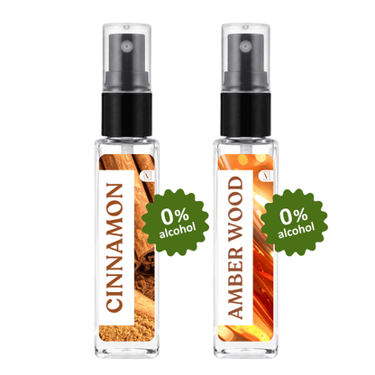 Two clear glass perfume spray bottles on a white background, labeled Cinnamon and Amber Wood, each with a black spray top and a green '0% alcohol' badge.