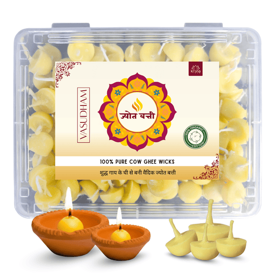 A clear plastic box of Vasudham 100% Pure Cow Ghee Wicks with a cream-colored label, shown alongside two lit clay diya lamps and several unlit yellow ghee wicks on a white background.