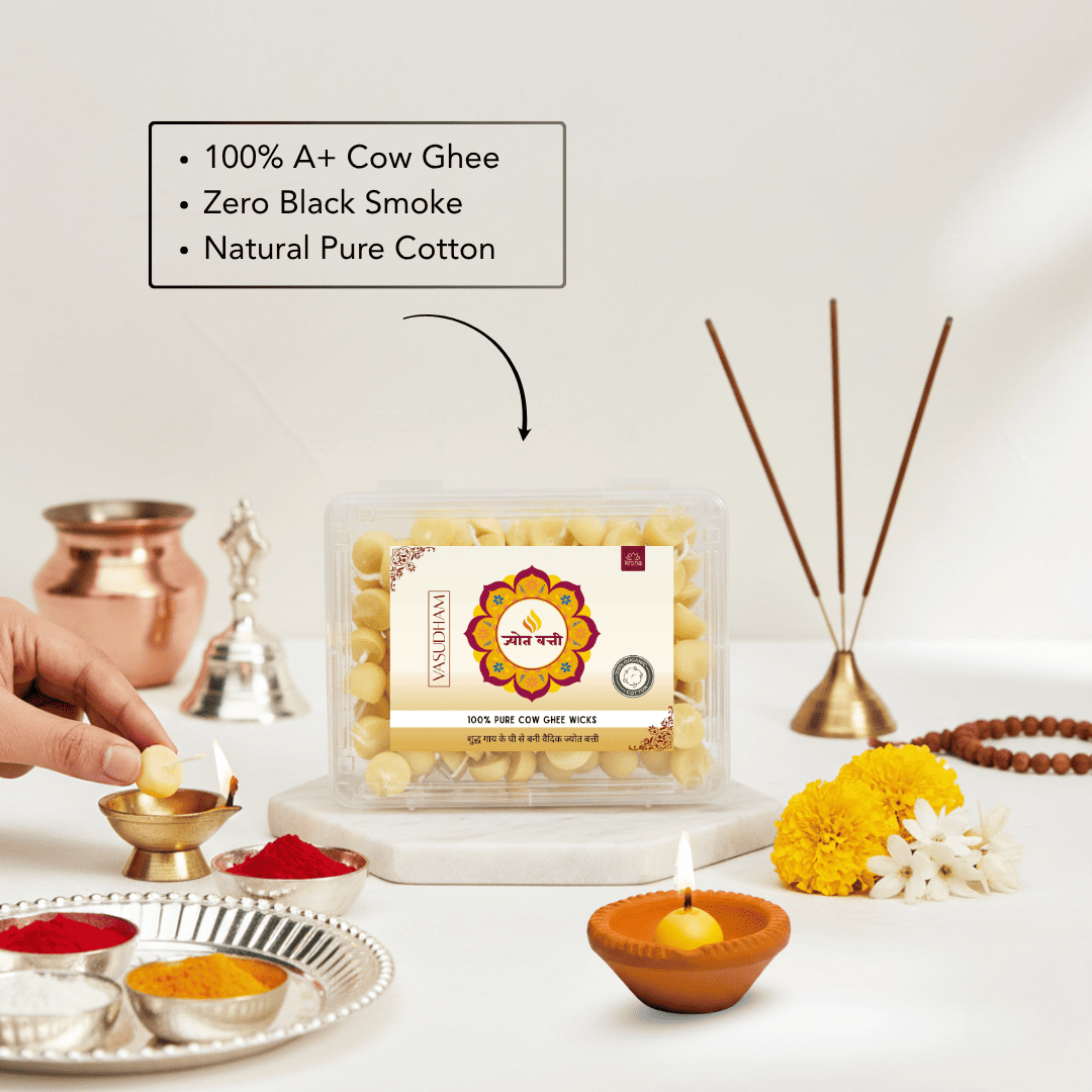A clear plastic tub of Vasudham 100% A+ Cow Ghee wicks on a white marble pedestal, surrounded by warm ceremonial props including brass incense sticks, a copper diya holder with a lit oil lamp, yellow marigolds, and a hand placing a small ghee lamp on a silver tray. A text box highlights product benefits: '100% A+ Cow Ghee', 'Zero Black Smoke', and 'Natural Pure Cotton'.