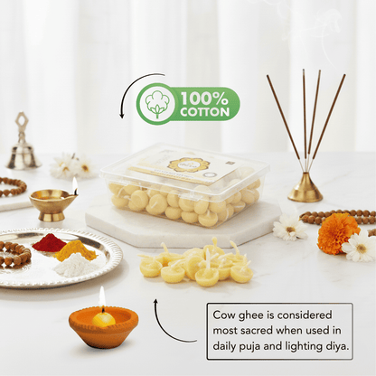 A styled product display featuring a clear plastic container of yellow wax diyas (oil lamps) on a white marble slab, surrounded by decorative props including brass diya lamps, incense sticks, marigold flowers, and a plate with colorful powders. Text overlays highlight '100% COTTON' and describe the spiritual significance of cow ghee for lighting diyas.