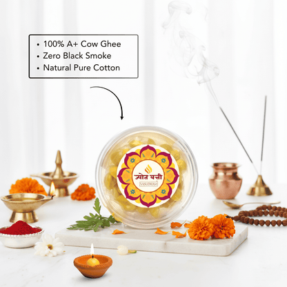 A clear round jar of Ghee Ganesha idol wax beads sits on a marble tray, surrounded by ritual props including brass oil lamps, marigolds, incense sticks, and a text callout emphasizing 100% A+ Cow Ghee, Zero Black Smoke, and Natural Pure Cotton.