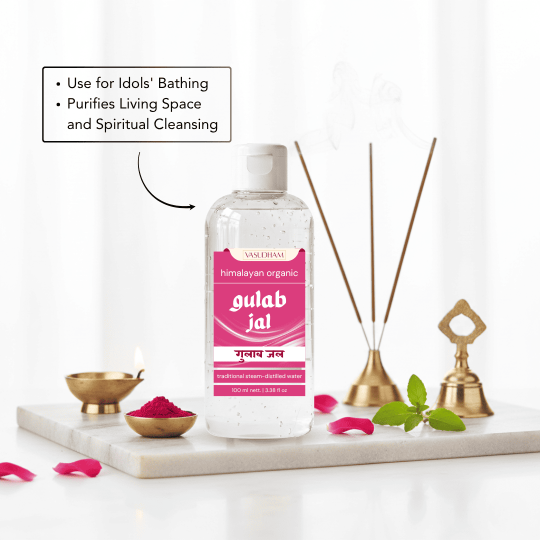 A bottle of Vasudham Himalayan organic spiritual cleansing rose water liquid stands on a white marble tray with incense sticks, brass lamps, green leaves, and pink petals. A text box on the left reads, 'Use for Idols' Bathing', 'Purifies Living Space and Spiritual Cleansing'.