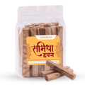 A clear stand-up pouch containing wooden incense sticks, with a yellow front label reading 'VASUDHAM' and Devanagari text, plus several loose samidha sticks placed in front on a white background.