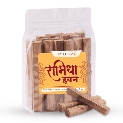 A clear stand-up pouch containing wooden incense sticks, with a yellow front label reading 'VASUDHAM' and Devanagari text, plus several loose samidha sticks placed in front on a white background.