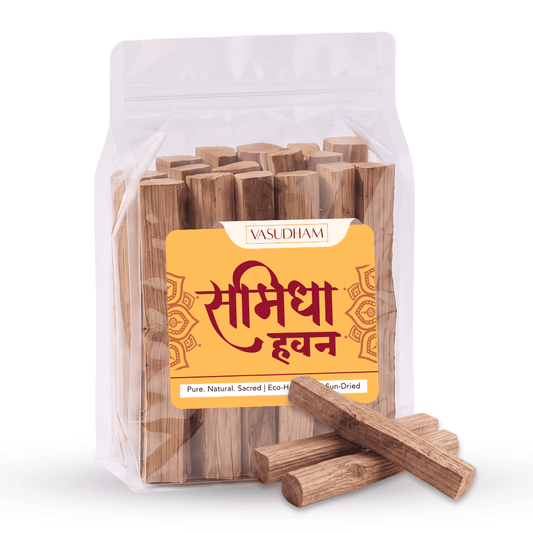 A clear stand-up pouch containing wooden incense sticks, with a yellow front label reading 'VASUDHAM' and Devanagari text, plus several loose samidha sticks placed in front on a white background.