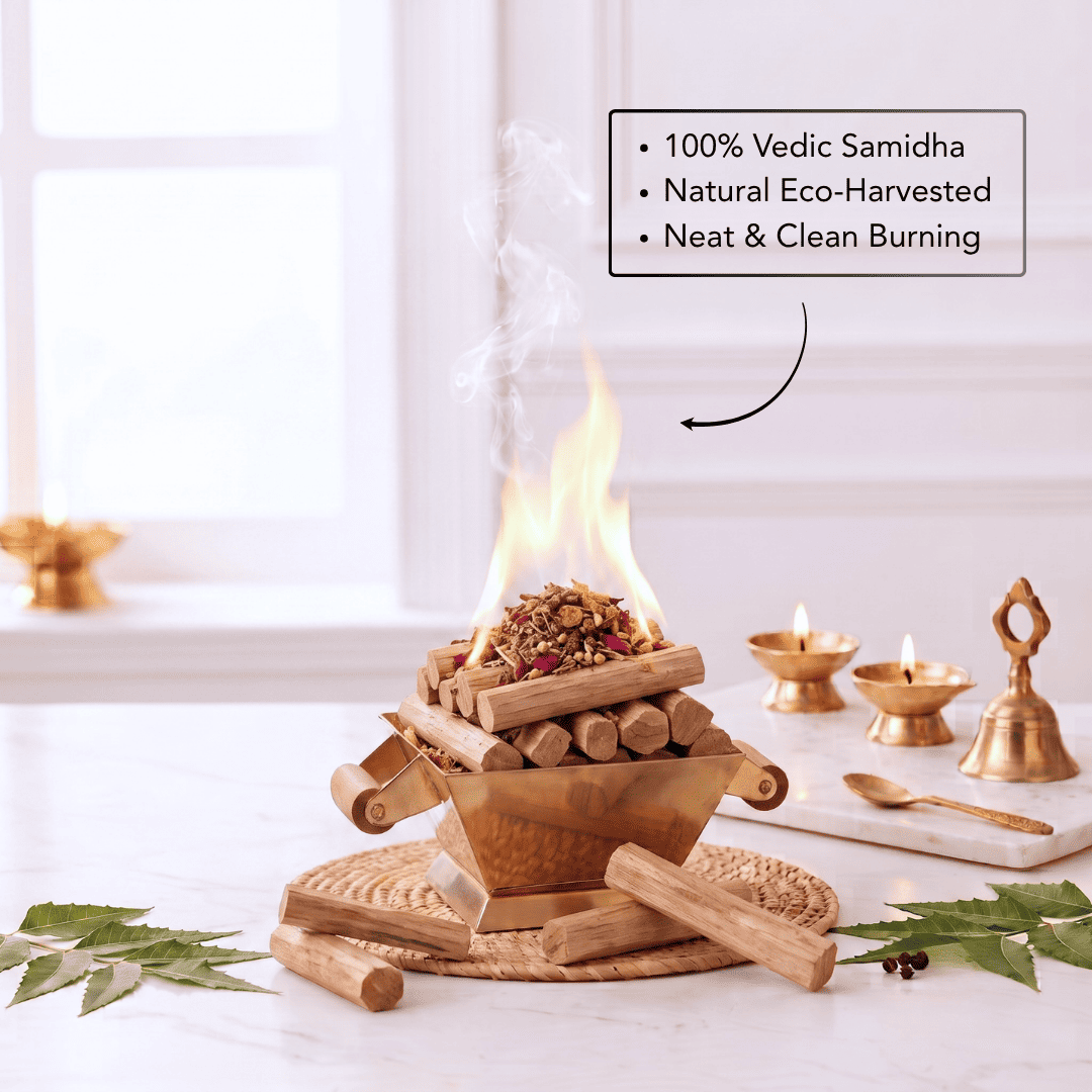 A small wooden burning vessel with visible flames sits on a tabletop in a bright indoor setting. The scene includes additional brass oil lamps, a spoon, leaves, and a text callout describing the product as 100% Vedic Samidha, natural eco-harvested, and neat clean burning.