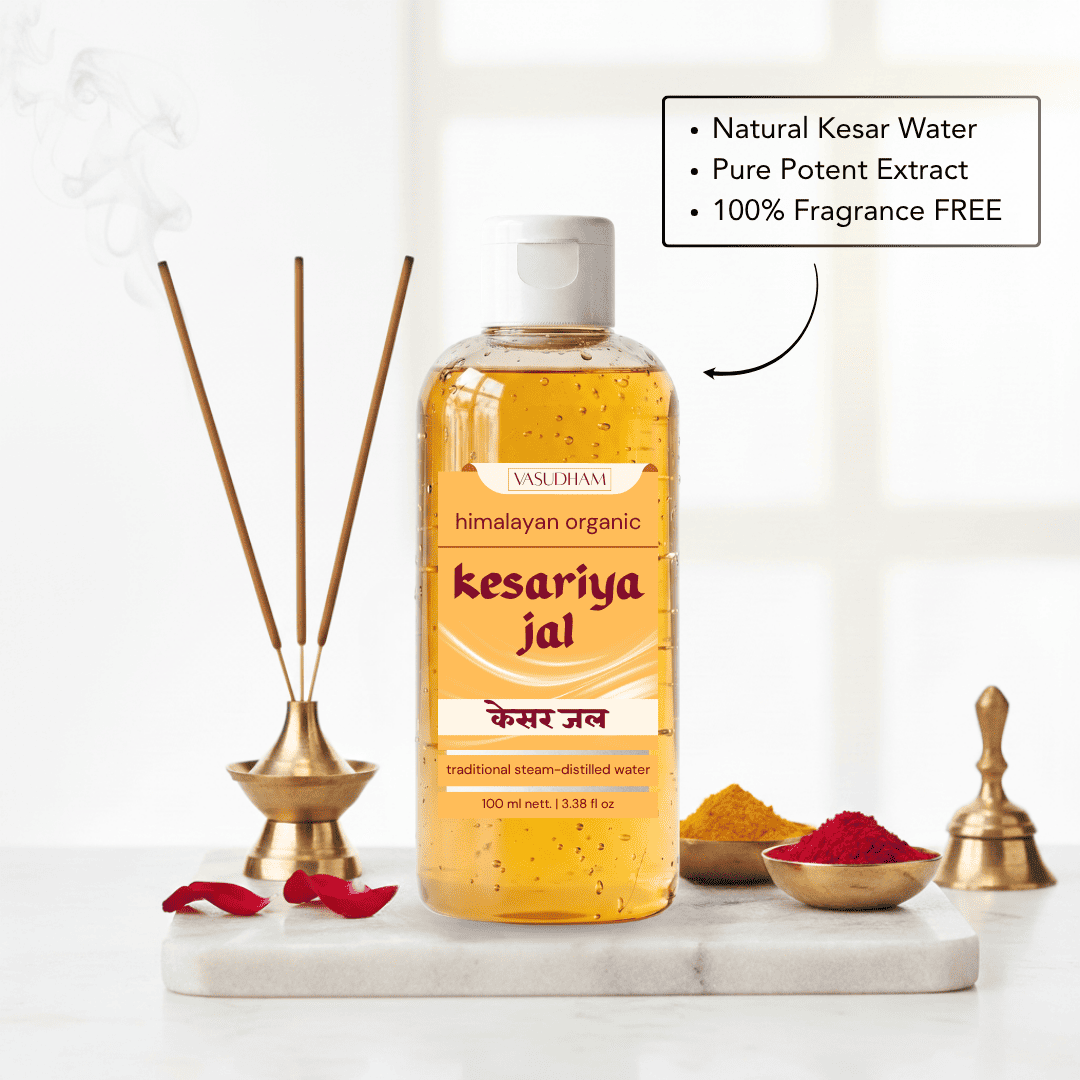 A bottle of Vasudham 'Kesariya Jal' traditional steam-distilled water, shown on a white marble tray with incense sticks, brass bowls of spices, and a brass bell against a bright, softly blurred background. A text box highlights key product features.