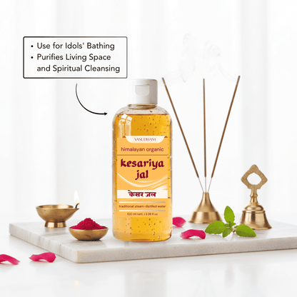 A bottle of Vasudham Kesariya Jal (Himalayan Organic Sacred Floral Water) displayed on a white marble tray with decorative bath accessories, pink petals, and a text box explaining its benefits.