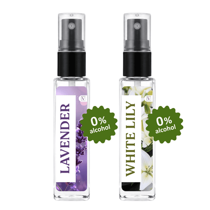 Two clear glass perfume atomizer spray bottles with black spray tops, shown side by side on a white background. The left bottle has a lavender floral label and the right bottle has a white lily floral label, both with green starburst badges reading '0% alcohol'.