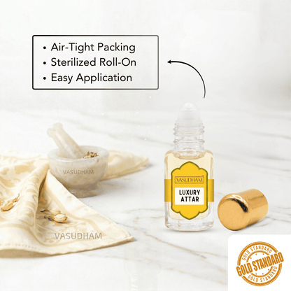 A clear glass roll-on bottle labeled 'VASUDHAM LUXURY ATTAR' with a yellow label and gold cap, shown on a light marble surface with a gold cradle and a folded cream cloth. Text highlights air-tight packing, sterilized roll-on, and easy application.