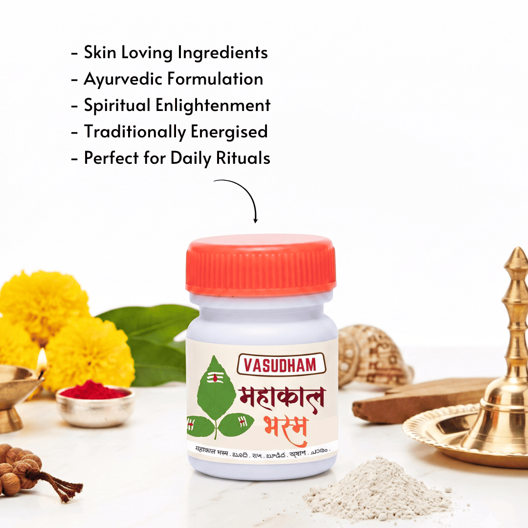 A white plastic bottle of Vasudham Mahakal Mantra powder with a bright orange-red cap, displayed on a white background with Ayurvedic props like yellow marigolds, a brass lamp, and powder. Text on the top left lists product benefits in English.