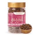 A jar of Vasudham Indian Guggul herbal powder with a gold lid and a pink label, shown alongside a small wooden bowl filled with the dark brown powder on a white background.