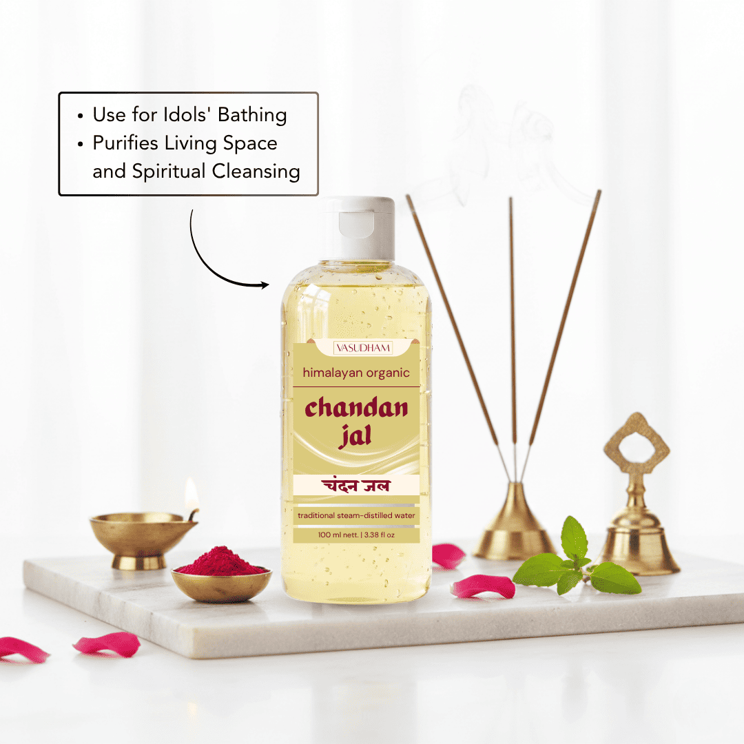 A bottle of Vasudham Chandan Jal traditional cleansing oil stands on a white marble tray with decorative brass oil lamps, reed diffuser sticks, green leaves, and pink petals. A text callout on the left reads 'Use for Idols' Bathing' and 'Purifies Living Space and Spiritual Cleansing'.