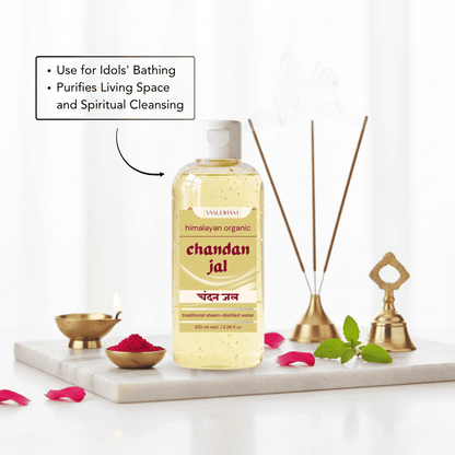 A bottle of Vasudham Chandan Jal traditional cleansing oil stands on a white marble tray with decorative brass oil lamps, reed diffuser sticks, green leaves, and pink petals. A text callout on the left reads 'Use for Idols' Bathing' and 'Purifies Living Space and Spiritual Cleansing'.